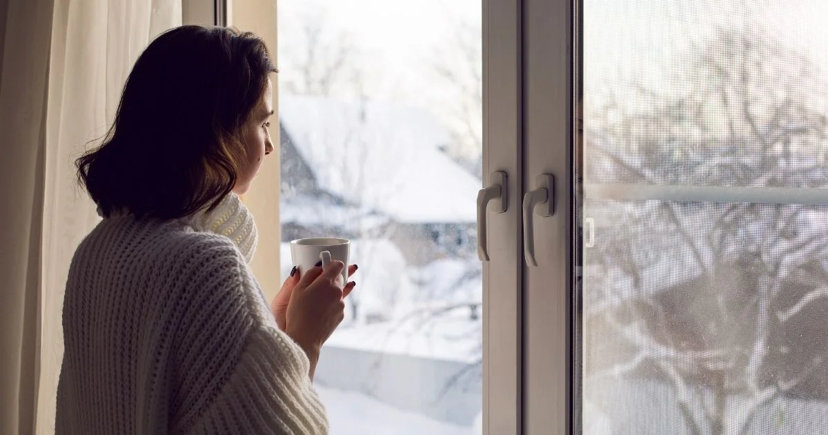 Woman in cozy sweater holding a mug and looking out snowy window.